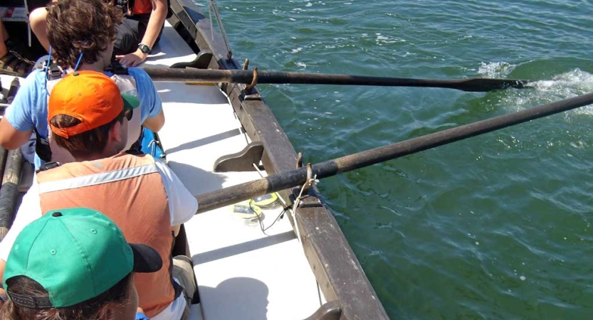 A group of students maneuver a sailboat using oars. 